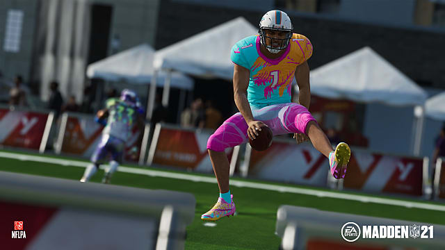 A player in an orange and teal jersey, blue and purple pants, hurdles with ball in his hand on a training field.